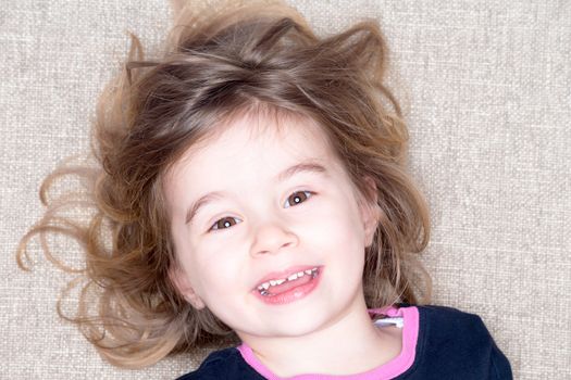 Pretty vivacious three year old little girl lying on a carpet with her hair flying around her face smiling happily up at the camera with a joyful expression