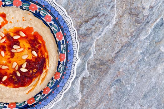 Bowl of delicious creamy homemade hummus made with mashed chickpeas, tahini, oil, lemon juice, and garlic, on a natural stone countertop with decorative blue pattern and copyspace