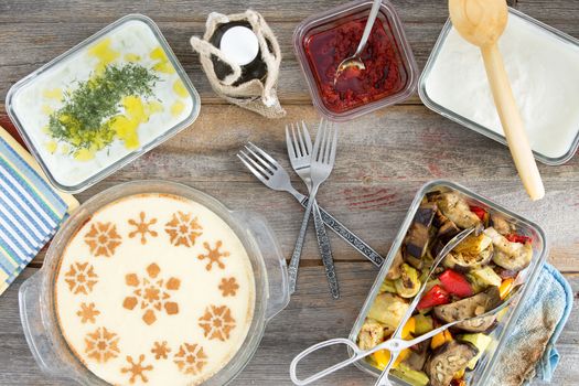 Delicious picnic lunch with grilled vegetables, tzatiki, yogurt with dill, pepper paste and a decorative milky semolina dessert , overhead view on a wooden table