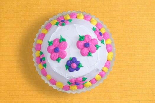Top view of a decorative iced homemade cake with colorful pink and blue flowers surrounded with a yellow and pink alternating border, served uncut on a yellow background with copyspace
