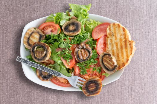 Grilled mushroom salad with fresh frilly lettuce and tomato served with toasted bread for a healthy lunchtime snack, viewed from above on a beige textile