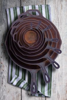 Collection of round rusty cast iron frying pans in diminishing sizes stacked one inside the other, viewed from above on a rustic napkin on an old wooden table