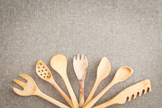 Fanned display of a variety of different wooden kitchen utensils arranged centered at the bottom over a neutral textile background with copyspace