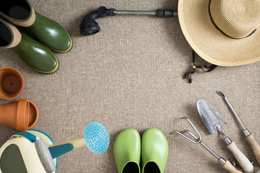 Border or frame of gardening tools on a neutral beige textile background with garden shoes and boots, sunhat, watering can, hand tools and flowerpots, with central copyspace, view from above