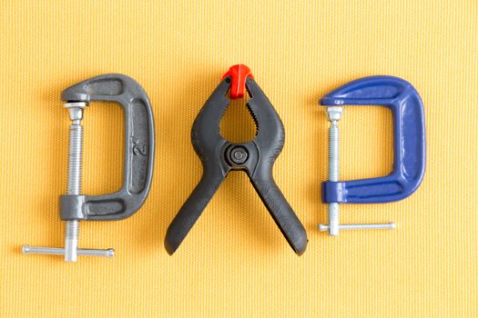 Assorted clamps from Dad's tool kit with adjustable grey and blue G-clamps and a spring loaded plastic A-clamp arranged in a line on a yellow background in a DIY concept