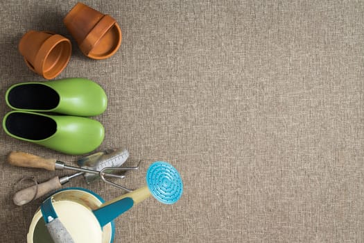 Side border of gardening tools and equipment including green garden shoes, flowerpots, hand tools and a watering can arranged in a line, overhead view with copyspace on a beige textile