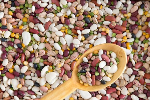 Background of 15 assorted beans and legumes with a spoonful in a wooden spoon including kidney, lima, black-eyed, yellow eye, garbanzo and pinto beans, lentils, split peas, overhead view in a layer