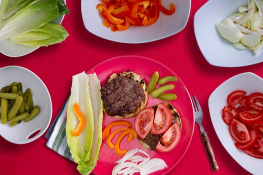 Creating the perfect healthy hamburger with two grilled beef patties on buns with sliced fresh ingredients including sweet pepper, dill cucumbers, tomato lettuce and onion on a red background