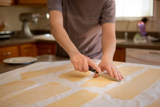 Hands of a man preparing fresh homemade fettuccine pasta trimming the sheets of rolled dough into rectangles to feed through the cutter on a table in a domestic kitchen