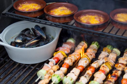 Delicious seafood outdoor meal at the BBQ with grilled salmon shish kebabs, marine mussels and individual pots of sausage stew, close up view on the grill