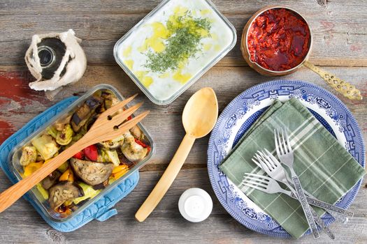 Grilled vegetables with eggplant, tzatziki, Turkish cacik with dill, and hot red chili pepper paste ready to be served with plates and cutlery on an old weathered wooden table