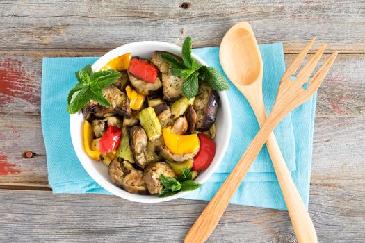 Healthy bowl of succulent Turkish grilled vegetables with eggplant, zucchini, red and yellow bell peppers and fresh sprigs of mint on a blue napkin on a rustic wooden table