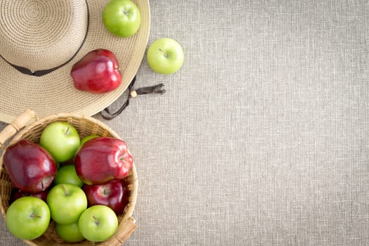 Farm fresh red and green apples direct from the farmer in a basket and on a wide brimmed straw sunhat viewed from above on a beige textile background with copyspace for a healthy diet