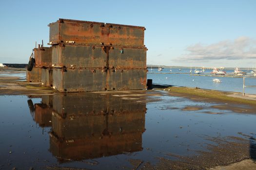 A stack of metal pontoons on the quayside with a marina and moorings offshore.