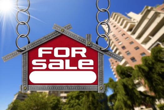 Grey metallic meter ruler in the shape of house with text for sale. For sale real estate sign with tall and blurred buildings in the background