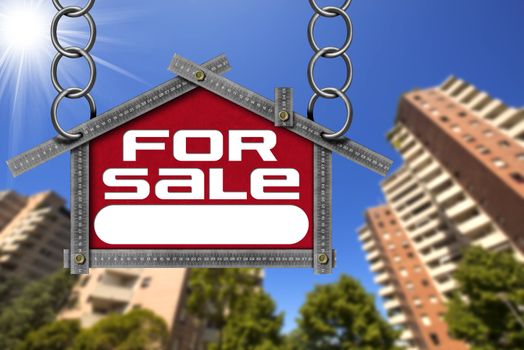 Grey metallic meter ruler in the shape of house with text for sale. For sale real estate sign with tall and blurred buildings in the background