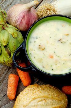 Healthy Artichoke Creamy Soup with Leek, Carrot, Garlic and Cheese in Dark Blue Pannikin with Sesame Bun closeup on Rustic Wooden background. Top View