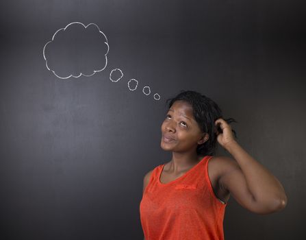 South African or African American woman teacher or student thinking, scratching her head standing against a blackboard background with chalk thought clouds