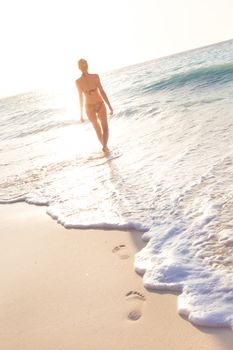 Happy woman enjoying in summer, walking joyfully on tropical beach in sunset. Beautiful caucasian model wearing bikini on vacations on sandy beach. Footprints in sand. 