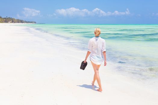 Caucasian woman walking joyfully on tropical beach. Beautiful caucasian model  wearing white beach tunic on vacations walking down picture perfect Paje beach, Zanzibar, Tanzania. Copy space.