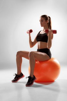 Sporty woman doing aerobic exercise with red dumbbells on a fitness ball on grey background