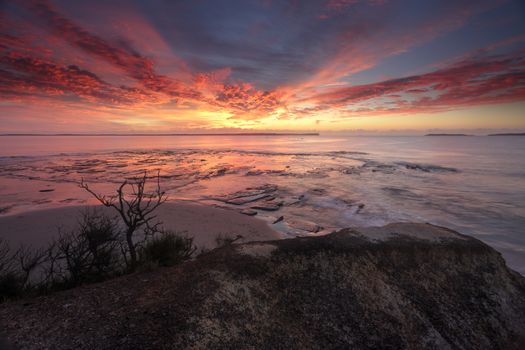 Spectacular sunrise over Plantation Point Vincentia.  Views from a rock ledge overlooking the reef at a high tide  and beyond to Beecroft Peninsula and Bowen Island on the horizon.  The brilliant colours in the sky also reflecting into the bay