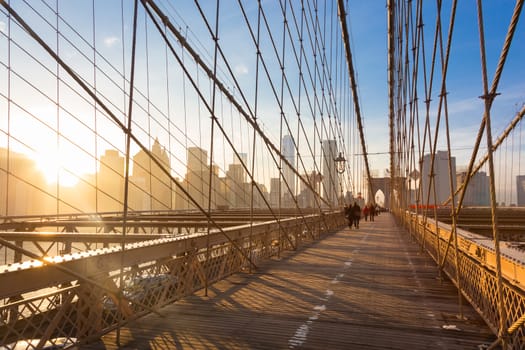 Couple walking on pedestrian path across Brooklyn bridge. New York City Manhattan downtown skyline in sunset with skyscrapers illuminated over East River panorama as seen from Brooklyn bridge.