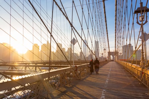 Couple walking on pedestrian path across Brooklyn bridge. New York City Manhattan downtown skyline in sunset with skyscrapers illuminated over East River panorama as seen from Brooklyn bridge.