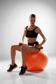 Sporty woman doing aerobic exercise with red dumbbells on a fitness ball on grey background