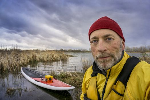 senior paddler in life jacket with his paddleboard and lake in background, early spring scenery with stormy sky in Colorado
