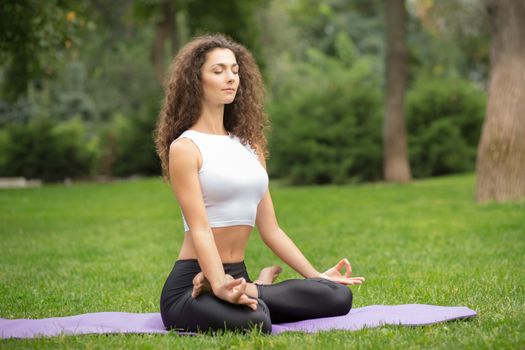 Pretty woman with eyes closed doing yoga meditation in the lotus position. Green grass background 