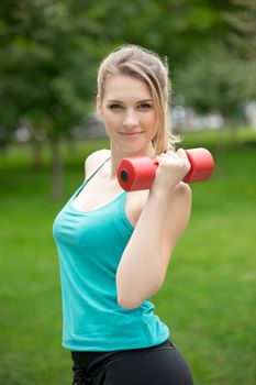 Young beautiful sports girl with dumbbells in the park on a background of green grass