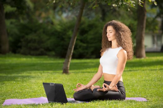 Meditating sporty young woman with laptop. Green grass background 
