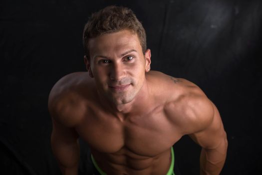 Handsome shirtless bodybuilder shot from above, standing on dark background wearing trunks