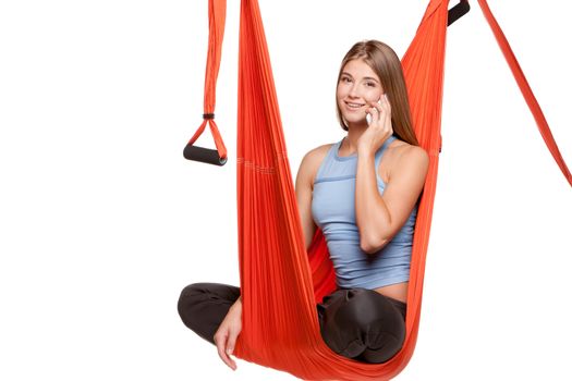 Young woman sitting in red hammock for anti-gravity aerial yoga with phone on a white background.