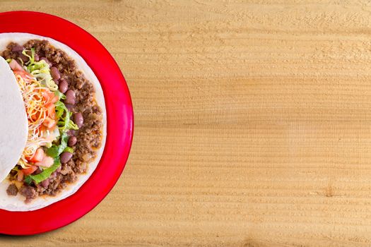 Delicious burrito filled with spicy ground beef , beans and salad topped with cheese on a colorful red plate, overhead view on a wood background with copyspace
