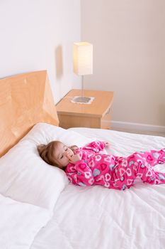 Adorable small girl in pink pajamas lying resting on a big king size bed picking her nose as she awakens from her daily nap
