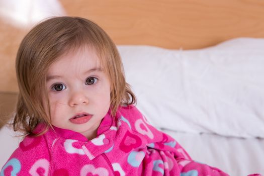 Cute young girl with tousled hair wearing pink pyjamas just awoken from her sleep giving the camera a wide eyed bemused stare