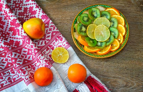 On the table is a dish of sliced fruits: oranges, kiwi, lemon. Near them on a beautiful towel are two oranges and an Apple.