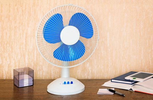 On the table is a handy fan with switching speeds , books, writing materials.