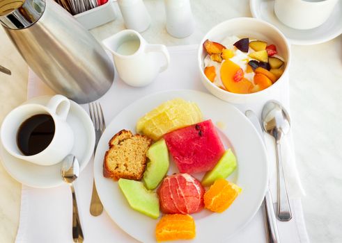 On the white tablecloth is a plate with a variety of fruits, standing next to a bowl of cream and peaches, as well as a coffee pot and a Cup of black coffee.