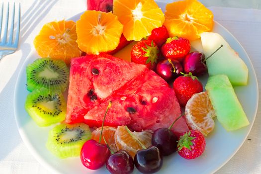 Strawberries, cherries, slices of watermelon, slices of Mandarin are located on a plate on a white tablecloth table.