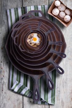 Sunny side up fried egg and mushrooms in a nested stack of old cast iron frying pans in a rustic kitchen, overhead view