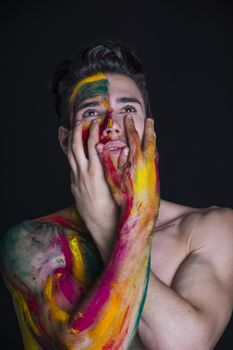 Attractive young man shirtless, skin painted all over with bright Holi colors on dark background