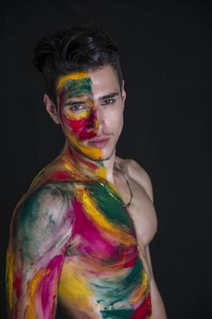 Attractive young man shirtless, skin painted all over with bright Holi colors on dark background