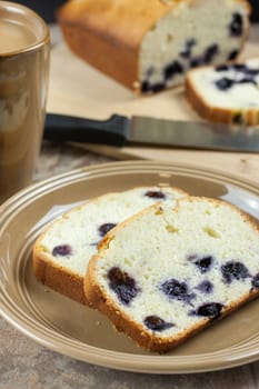 Sliced lemon blueberry bread on a plate with a cup of coffee and the remaining loaf in the background.