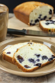Sliced lemon blueberry bread on a plate with a cup of coffee and the remaining loaf in the background.