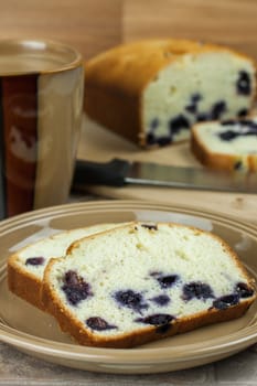 Sliced lemon blueberry bread on a plate with a cup of coffee and the remaining loaf in the background.