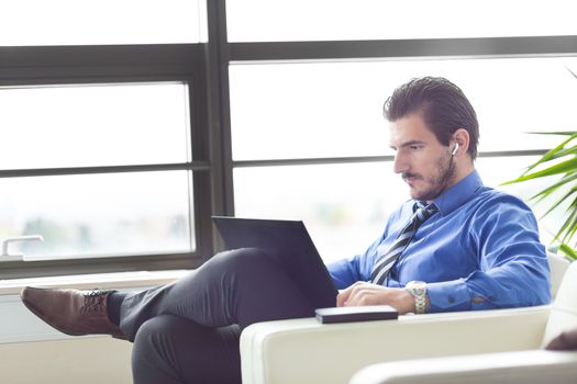 Busy, focused businessman in office working on his laptop wearing headphones. Side view. 