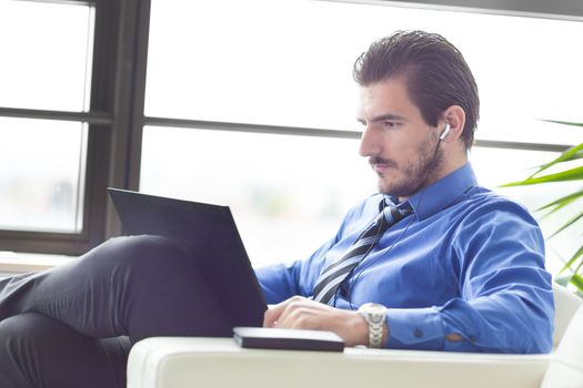 Busy, focused businessman in office working on his laptop wearing headphones. Side view. 